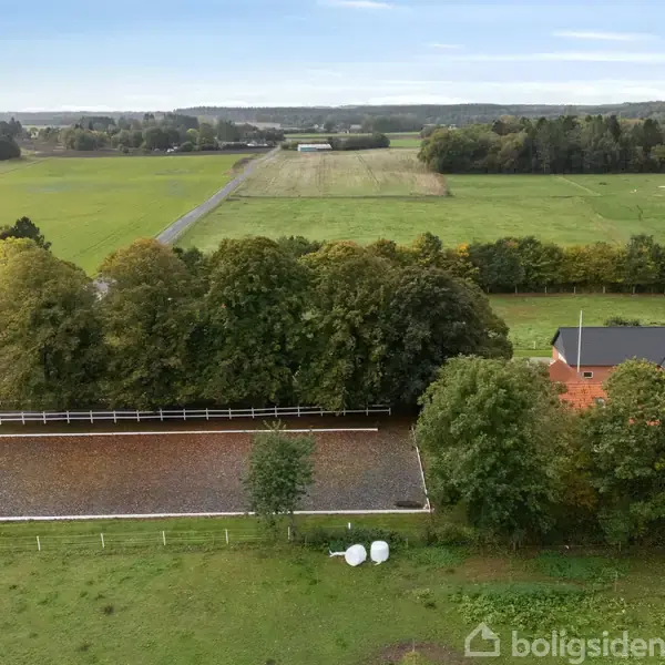 En stor grøn mark omkranser en indhegnet ridebane. En rød tagbygning er synlig ved siden af træer. Landskabet strækker sig i horisonten under en blå himmel.