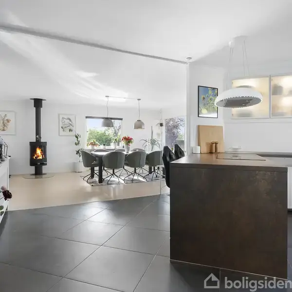 Pejs står brandende i hjørnet af et moderne, åbent køkken-alrum med spisebord og stole. Væggene er lyse, og der ses indretningsdetaljer som malerier og en gulvplante.