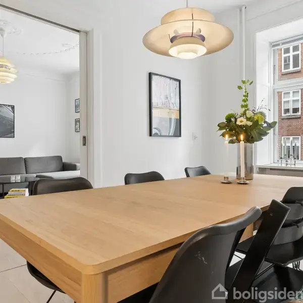 Et træbord med sorte stole omkring står i et spisestueområde ved siden af en stor vindueskarm med planter. En sofa kan ses i det tilstødende værelse.
