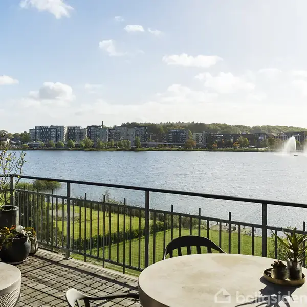 Balkon med runde borde og planter set fra en høj bygning. Udsigt over sø med springvand, moderne bygninger og kraner i baggrunden. Solskin og blå himmel skaber en lys atmosfære.