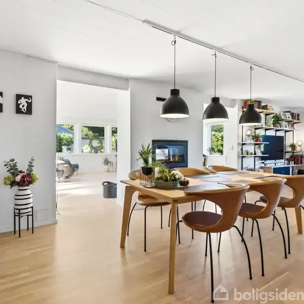 Et spisebord med moderne stole står i et lyst rum. Der hænger lamper over bordet, og i baggrunden ses en reol, planter og store vinduer med udsigt til natur.