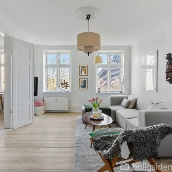 Stue med en grå sofa og sofabord med blomster i vase, set mod store vinduer og åbne døre til et tilstødende rum. Væggene er dekoreret med kunst og en maske.