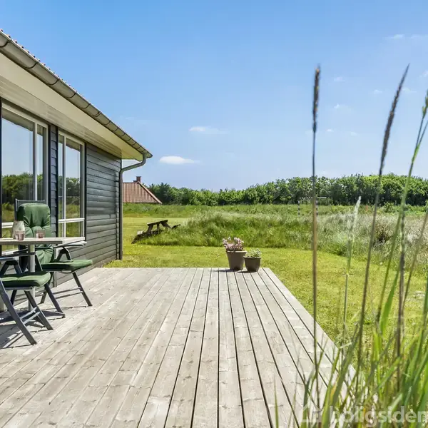 Træterrasse med to grønne stole og et lille bord ved siden af et sortmalet hus, omgivet af en græsplæne og marker i solskin. En bænk ses i baggrunden.