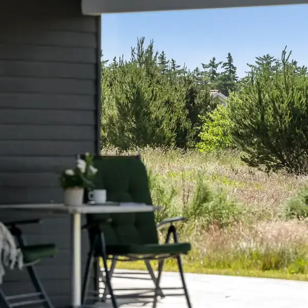 To grønne stole og et bord med en blomst og krus står på en terrasse, omgivet af græs og buske i et naturpræget landskab.