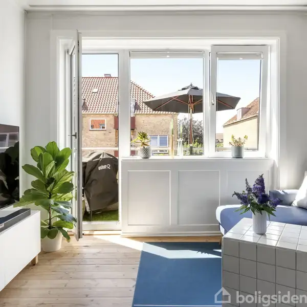 Stue med blå sofa og hvidt sofabord, plante i hjørnet. Stor skydedør åben til altan med udsigt til hus og himmel. Fladskærms-TV på skænk til venstre.