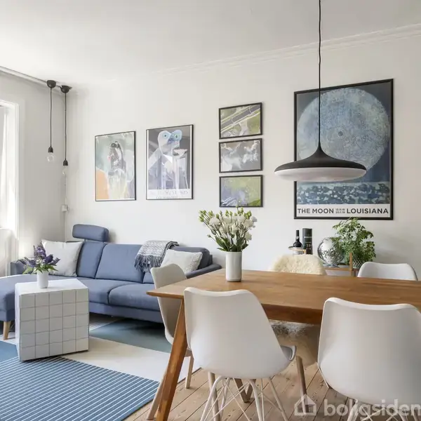 Stue med blå sofa ved vindue, træbord med fire hvide stole, plakater på væggen. En lampe hænger over bordet. Tekst på plakat: "THE MOON OVER LOUISIANA".