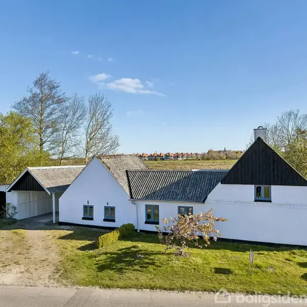 Hus med hvid facade og mørkt tag står på grøn græsplæne, omgivet af træer. Skyfri blå himmel danner en rolig baggrund. Ingen tekst er synlig.
