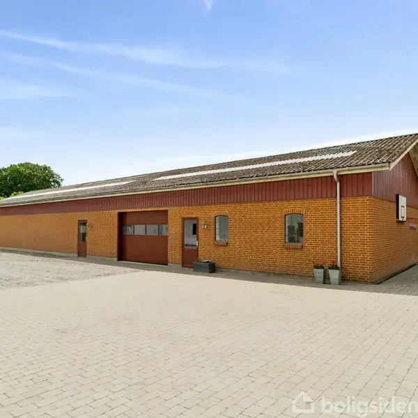 En stor, rektangulær bygning af røde mursten står på en flisebelagt grund. Omkring bygningen er der grønne buske og træer under en klar blå himmel.