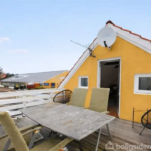 Et gult hus med rød tag har en udendørs terrasse, hvor der står et bord og flere stole. En parabolantenne er monteret på gavlen. Placeringen er i et beboelsesområde.