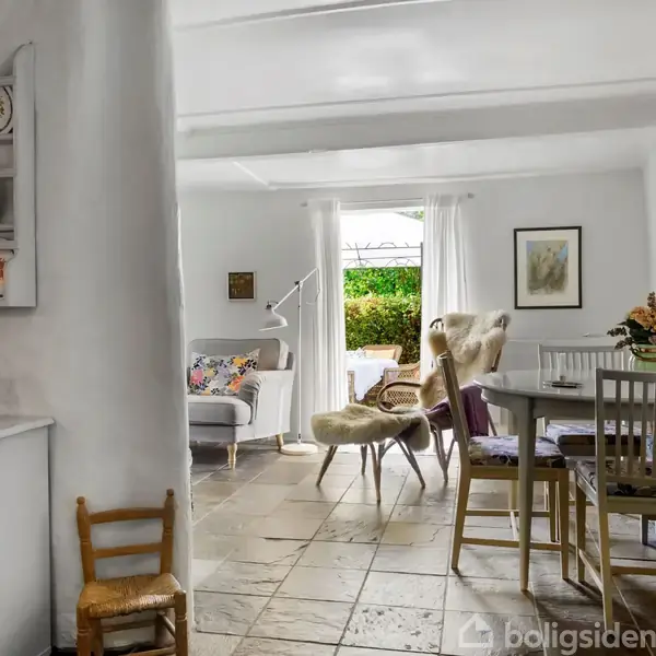 Stue med spisestue bord og stole; i baggrunden en sofa og lænestol med udsigt til en have gennem store vinduer. Væghylder med tallerkener til venstre.