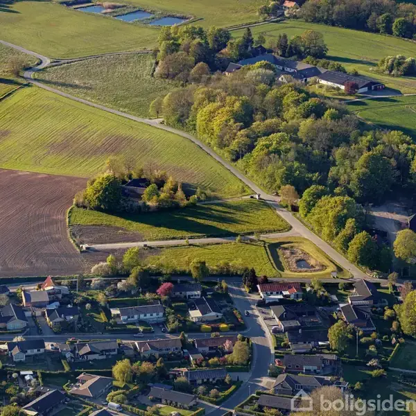 Luftfoto af et landskab, der viser en lille landsby med huse og veje, omgivet af marker og skove. Byen synes at være indlejret i et bakket, grønt område.