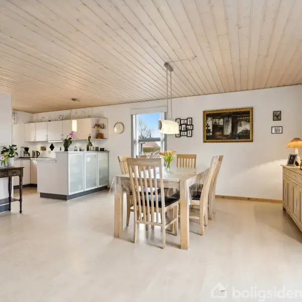 Et spisebord med seks stole står midt i rummets åbne stue-køkkenområde. Bordet er pyntet med en vase med blomster. Der er lyse trælofter og vægge dekoreret med billeder.