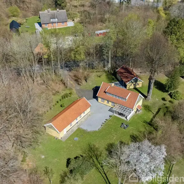 To huse med røde tage er placeret blandt træer i et skovområde. Der er en græsplæne og en betonsti mellem bygningerne, der skaber en landlig atmosfære.