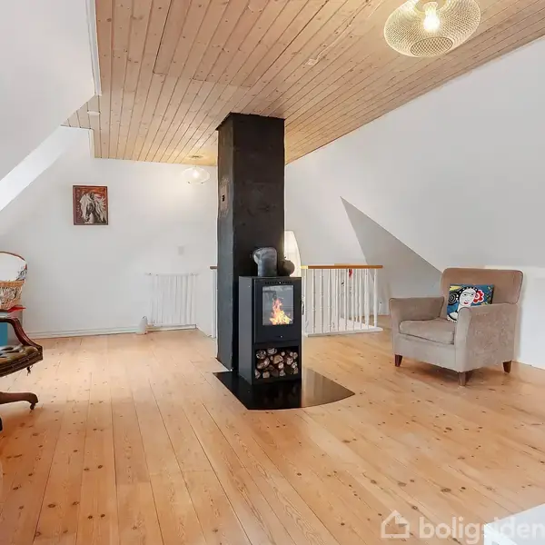 Brændeovn i midten af et lyst loftsrum med træpaneler. Omgivet af polstrede stole og en kurvestol, skaber en hyggelig stemning.