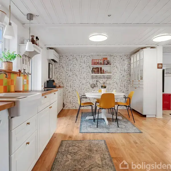 Køkken med hvide skabe, farverig mosaikvæg bag vasken. Et spisebord med fire gule stole står på et blåt tæppe. Et tapet med naturmotiv beklæder væggen.