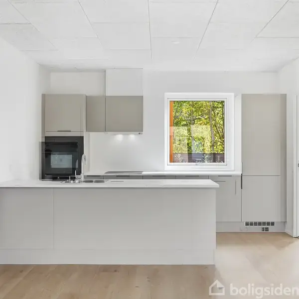 En moderne køkkenø med indbygget håndvask og komfur; ovn og opbevaringsskabe langs væggen; stort vindue og dør til højre, lysegråt gulv og hvide vægge og loft.