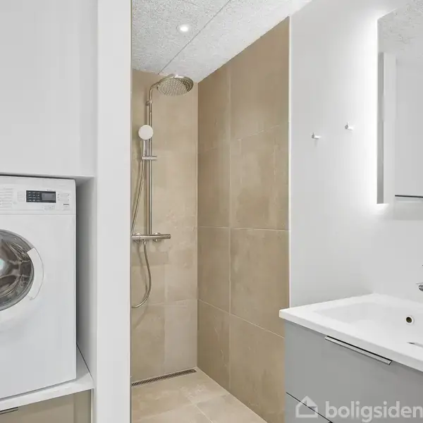 Vaskemaskine står i et lyst badeværelse med en åben brusekabine. Der er et moderne håndvaskarrangement med spejl på væggen. Atmosfæren er minimalistisk og funktionel.