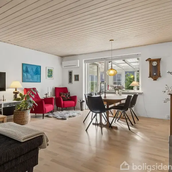 Stue med sofa, to røde lænestole og spisebord. TV på væggen. Store vinduer giver udsigt til en terrasse. Indrettet med lamper, et ur og kunst på væggene.