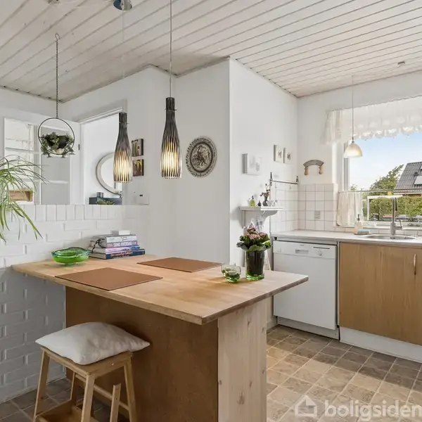 Et køkken med en træbordplade og barstol. Hængende lamper skinner over bordet. I baggrunden er et vindue med udsigt til en have. Planter og bøger pynter rummet.