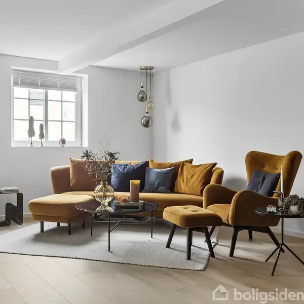 En moderne stue med en sennepsgul sofa og lænestol står på et gråt tæppe. Et sofabord er placeret foran dem, omgivet af minimalistisk indretning i lyse omgivelser.