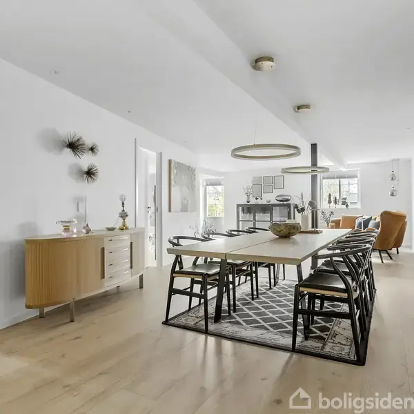 Et moderne, lyst spisestue med et langt spisebord omkranset af stole på et mønstret tæppe. Til venstre er en kommode og døre; i baggrunden ses en stue.