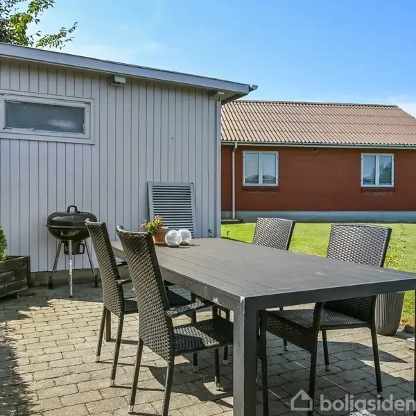 Sort havebord med seks stole står på en flisebelagt terrasse. I baggrunden ses et lille skur og en grill; et rødt hus er synligt over plænen under en klar, blå himmel.