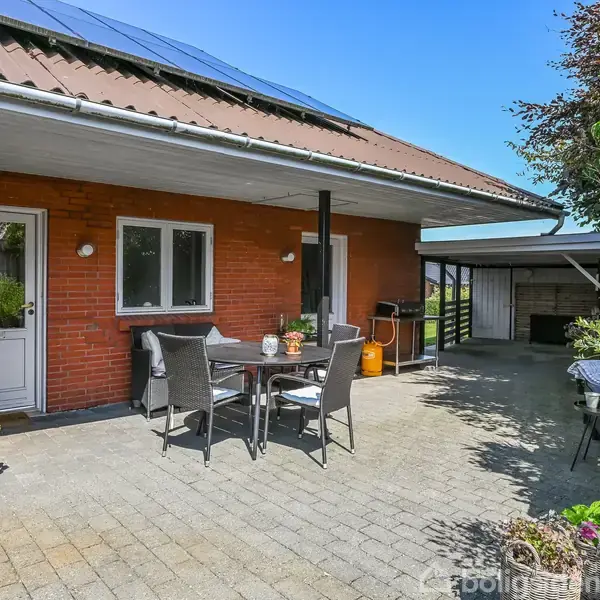 En terrasse med et bord og stole står foran et rødt murstenshus med en metaloverdækket tagrende. Til højre ses planter, en carport og en udendørs spiseplads.
