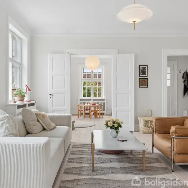 Stue med hvid sofa og brunt lædersofaarrangement omkring et sofabord med blomster. Store døråbninger fører til spisestue. Lyse vægge med moderat udsmykning giver et elegant udtryk.