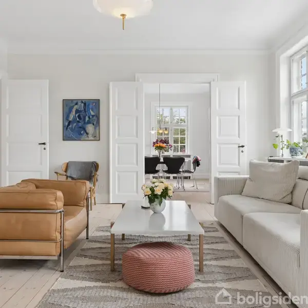 En lys stue med en hvid sofa og læderstol omringer et sofabord med blomster. To dobbeltdøre åbner til en spisestue med blomsterdekorationer. Vægkunst og store vinduer omkranser rummet.
