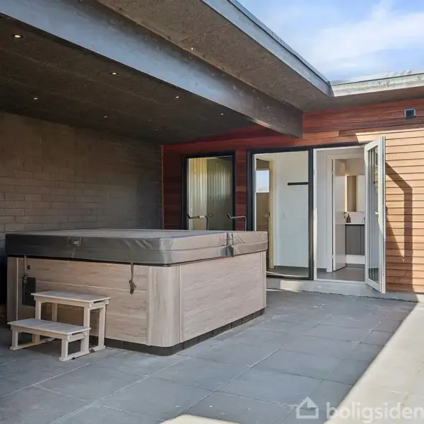 Udendørs boblebad står stille på en terrasse med grå fliser, beskyttet af et halvtag ved siden af et åbent dørparti af en moderne træbeklædt bygning.