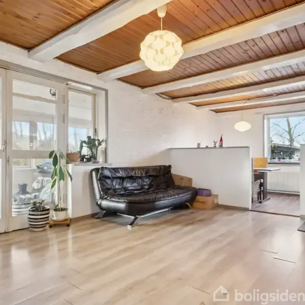 En stue med en sort lædersofa står tom. En brændeovn opvarmer rummet og et bord ses i baggrunden. Lyse trægulve og loftsbjælker skaber en hyggelig atmosfære.