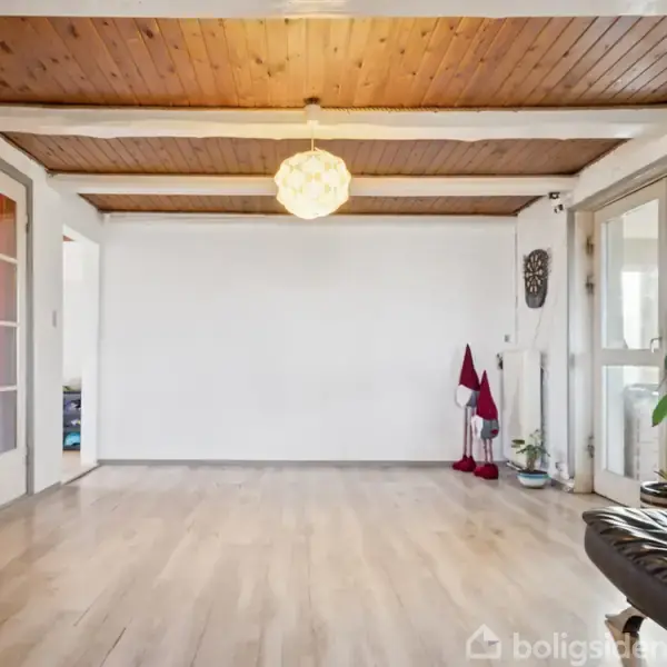 En tom stue med trægulv og træloft. En stor lampe hænger fra loftet. Der er åbne døre, planter og dekorationer i hjørnerne samt en mørk lænestol til højre.