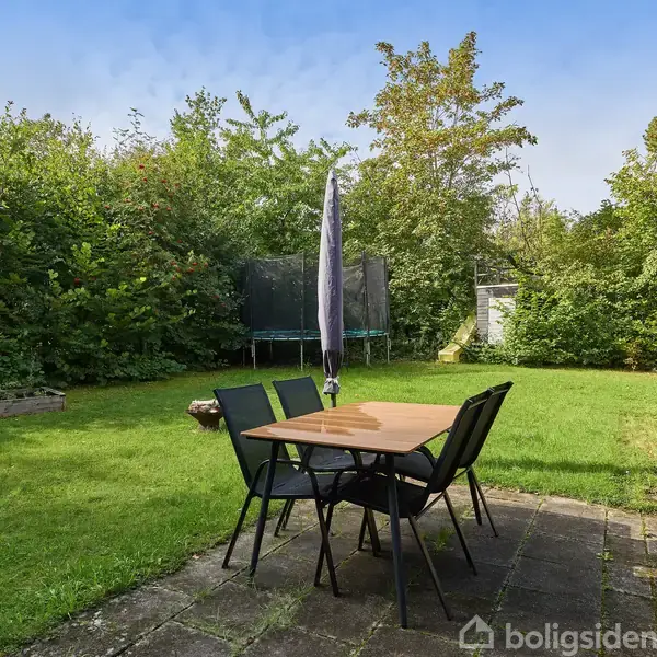 Et havebord med stole står på en flisebelagt terrasse. En parasol er lukket. En trampolin ses i baggrunden, omgivet af grønne buske og træer i en baghave.