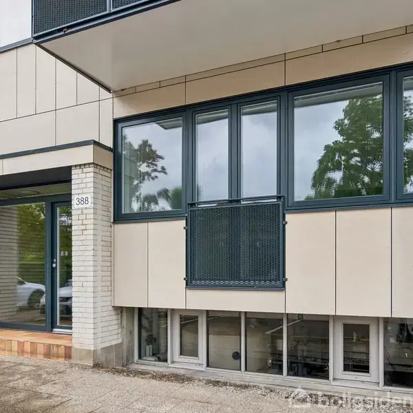 Bygning med beige facade og store vinduer. Indgangsdøren til venstre har husnummeret "388". En cykel står op ad væggen. Der er reflekser af træer i vinduerne.