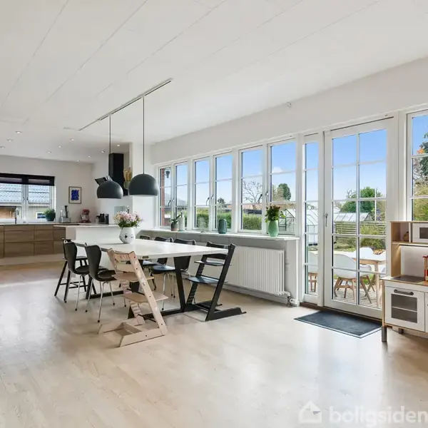 Spisebord med stole står i et lyst køkken-alrum med trægulv. Store vinduer giver udsigt til en have. En lille legekøkken står ved siden af en dør til haven.