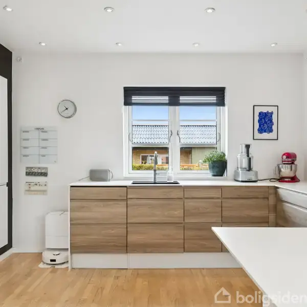 Moderne køkken med træskabsfronter og indbyggede hvidevarer. Et vindue med udsigt til tagene udenfor. Køkkenmaskiner står på bordpladen. Væggene er lyse, gulvet er af træ.