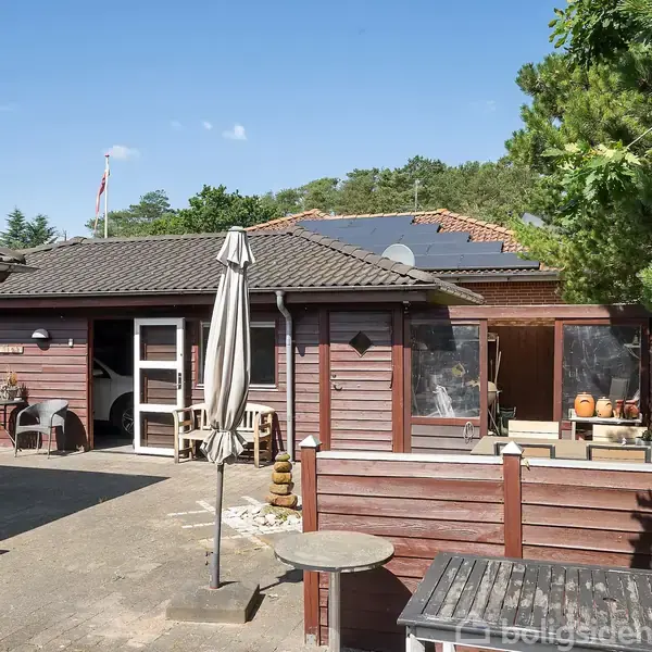 Et rødt træhus med solpaneler på taget står i en solrig gårdhave. En lukket parasol og havemøbler omkranser området, omgivet af grønne træer og en blå himmel.