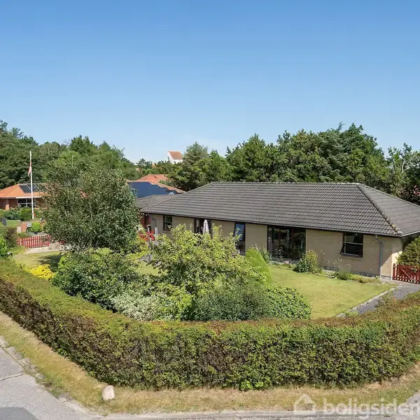 Et enfamiliehus med en gråtag, placeret i en frodig have, er omkranset af grønne hække og ligger i et fredeligt boligområde med veje og træer i baggrunden.