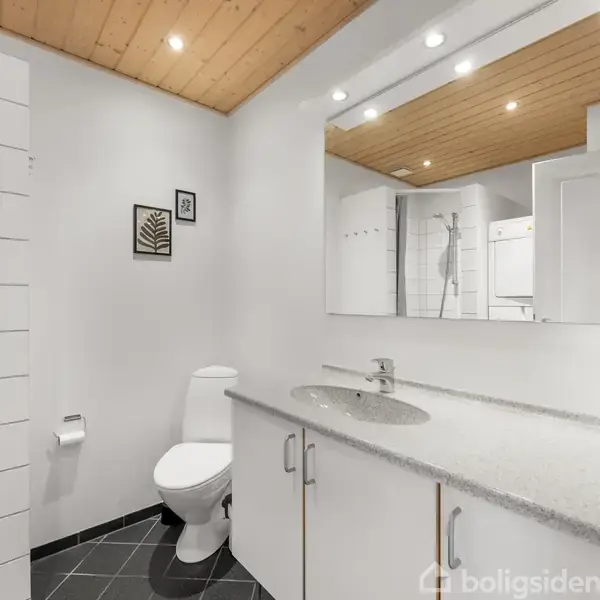 Et moderne badeværelse med toilet og håndvask på en grå bordplade. Et spejl med indbyggede spots reflekterer rummets træloft. Flisegulvet er mørkt, og der hænger billeder på væggen.