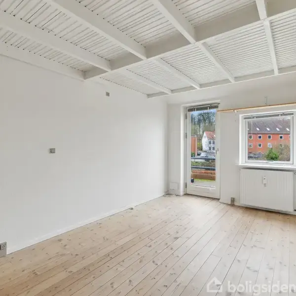 Et tomt rum med trægulv og hvidmalede vægge. Rummet har et vindue og en dør, der fører ud til en balkon. Udenfor ses nogle røde murstensbygninger.