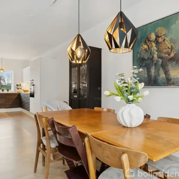 Et spisebord er dækket med en vase med blomster, omgivet af stole. Over bordet hænger dekorative lamper. I baggrunden ses et køkken og en væg med et maleri.