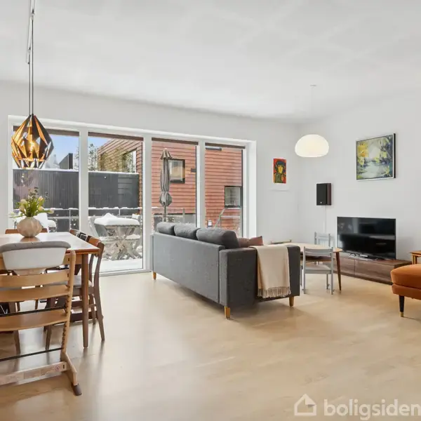 Stue med spisebord og stole ved siden af vinduer med udsigt til udendørsområde. En sofa og lænestol står overfor et tv. Billeder og lamper pynter væggene.