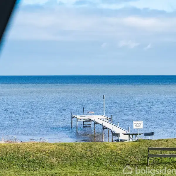 En træbro står stille og strækker sig ud i havet, omgivet af græs og en blå himmel. Et skilt ved broen indeholder tekst, men det er ulæseligt på billedet.