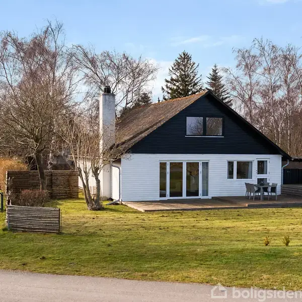 En hvidmalet villa med sort tag står på en grøn græsplæne. Villaen har en terrasse med havemøbler. Der er træer i baggrunden og gadenavnsskilte: "Skovstien" og "Mosevej".
