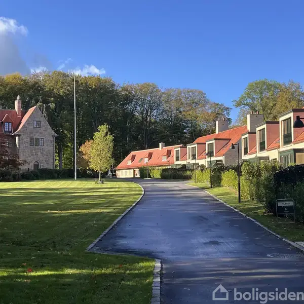 En række huse med rødt tag står langs en buet vej i et grønt område, omgivet af træer under en blå himmel. En større bygning ses til venstre.
