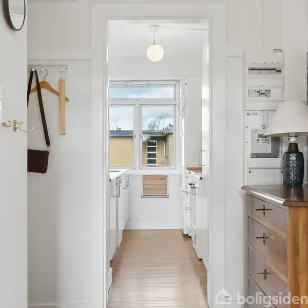 Gang med hvidmalede vægge fører til et lyst køkken. En trækommode med en bordlampe står til højre. Metalkroge med tøj hænger til venstre. Sollys kommer ind gennem vinduet.