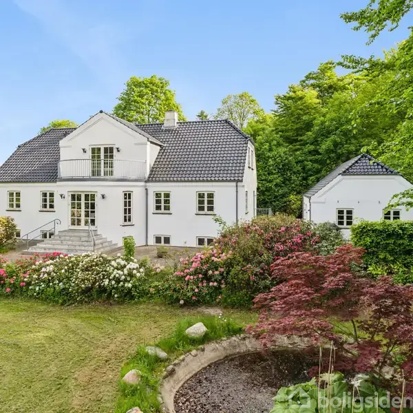 Hvidt hus med sorte tagsten står i en frodig have omringet af blomsterbede og grønne træer. Grussti fører op til hoveddøren.