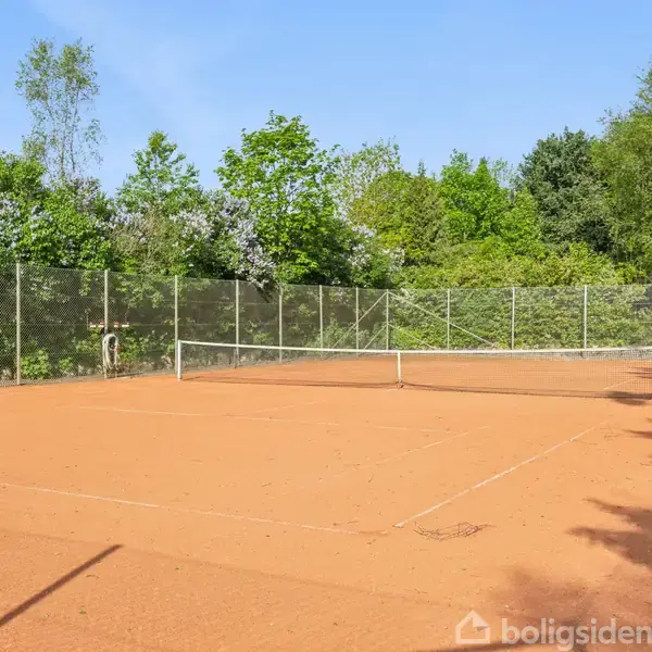 En tennisbane med grusbelægning står tom. Et net deler banen i to, omgivet af et hegn. Træer og grønne buske omkranser området under en blå himmel.