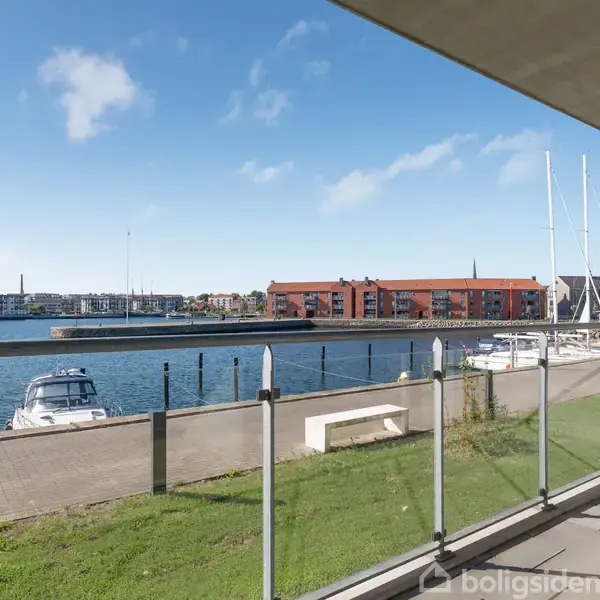 Balkon med glasrækværk tilbyder udsigt over en solbeskinnet marina, hvor både er fortøjet langs kajen. I baggrunden ses moderne bygninger og en klar blå himmel.