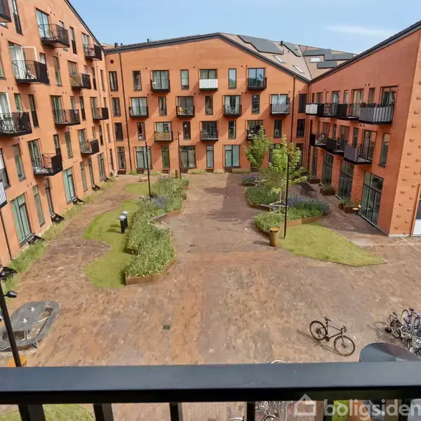 En rektangulær gårdsplads omgivet af moderne, orange murstensbygninger med altaner. Cykler står parkeret, og gården har stier og græsarealer med enkelte træer og bænke.